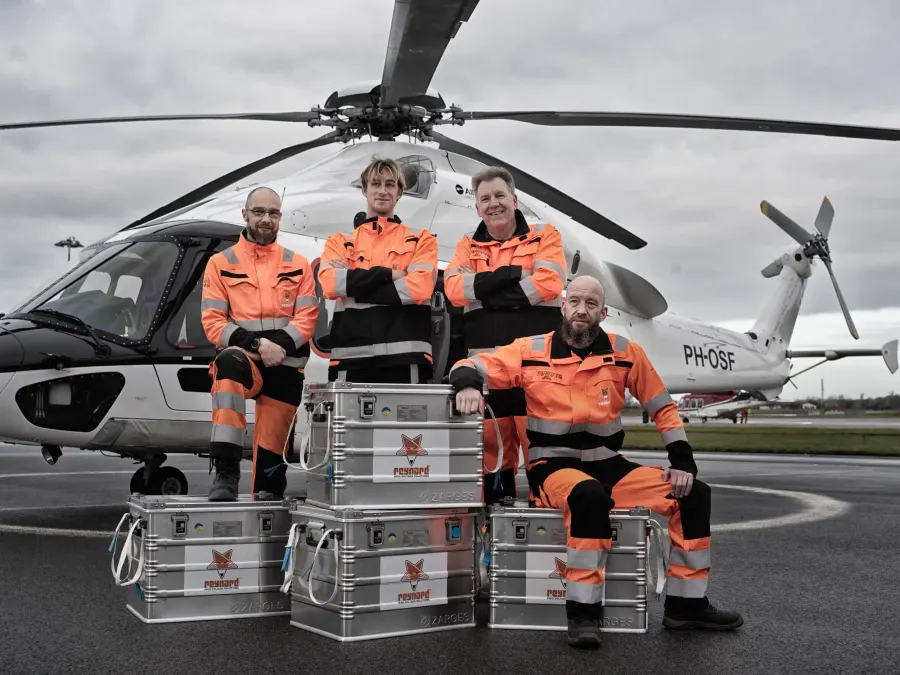 Four Reynard technicians in orange workwear in front of a helicopter, standing around aluminium tool cases marked with the Reynard fox